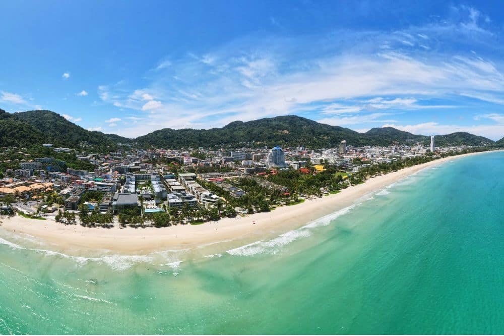 Aerial view of a coastal town with a long sandy beach adjacent to turquoise waters. The town features numerous buildings, including several taller structures, nestled among green hills. Patong nightlife comes alive beneath the mostly clear sky with a few scattered clouds. The shoreline curves gently along the coast—a true gem among the best places to visit in Patong.