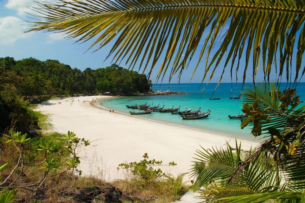 Tropical beach scene with a white sand shoreline, clear turquoise waters, and several long-tail boats anchored near the shore. Lush green palm trees and foliage frame the image, and gentle waves lap against the beach. The sky is bright blue with a few scattered clouds, hinting at nearby Patong Beach attractions.