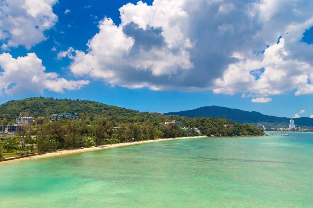 A scenic coastal view of a clear, turquoise ocean bordered by a sandy beach. Lush, green hills with scattered buildings rise in the background. White, fluffy clouds dot the vibrant blue sky, creating a serene and picturesque landscape—one of the best places to visit in Patong.