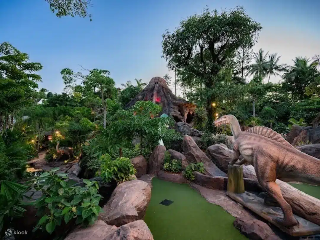 A lush, tropical mini-golf course features a dinosaur statue on the right. The course is surrounded by dense greenery, large rocks, and trees. In the background, a glowing volcano-like structure evokes a prehistoric jungle atmosphere under a clear twilight sky—perfect for your Phuket travel guide.