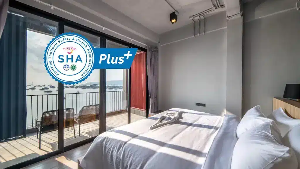 A modern hotel room with a large bed, crisp white linens, and a neatly folded towel. The room has floor-to-ceiling windows leading to a balcony with a view of the ocean and boats. The "SHA Plus" certification logo is overlaid in the top left corner, and Chalong Temple is just a short drive away.