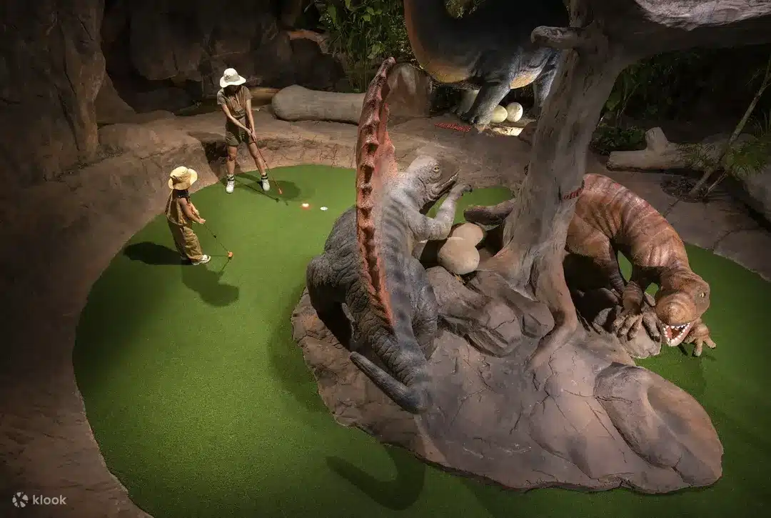 Two young children wearing safari hats play mini-golf on a green artificial turf surrounded by large, realistic dinosaur sculptures. One child is putting near a gray and red dinosaur, while another stands by a tree trunk. This captivating scene is one of the exciting Phuket attractions you'll find in any Phuket travel guide.