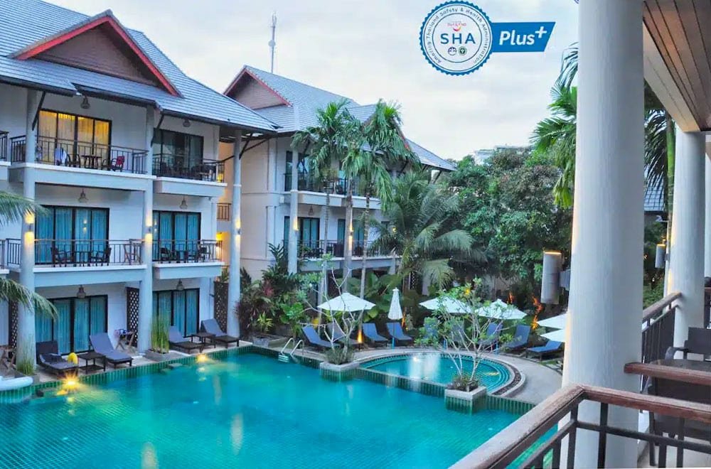 A serene resort view featuring two three-story buildings with balconies, surrounding a central turquoise swimming pool. The pool area includes lounging chairs, umbrellas, and lush tropical plants. The SHA Plus+ certification logo is displayed in the top right corner, indicating health and safety standards near Laem Phromthep Sunset Viewpoint.