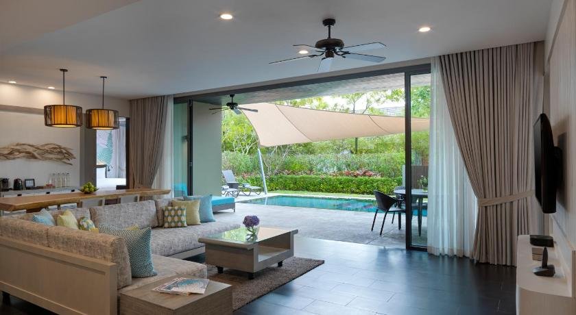 A modern, spacious living room with a large sliding glass door opens to a patio and pool area. Inside, there's a beige sectional sofa with colorful cushions, a wooden coffee table, and hanging pendant lights. Outside, the vibe mirrors popular Phuket attractions with outdoor seating overlooking a lush garden and inviting pool.