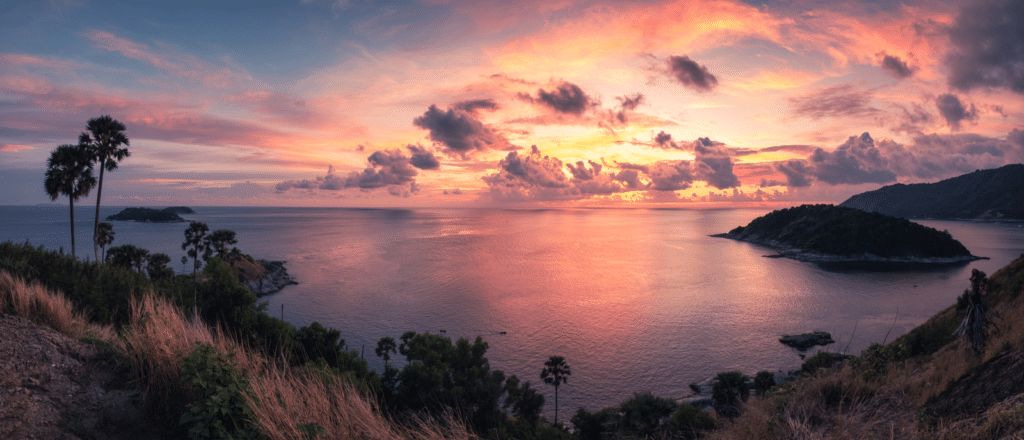 A stunning panorama of a sunset over a calm sea, with vibrant hues of orange, pink, and purple in the sky. Silhouetted island and mountains appear in the distance. A cliff with grasses, shrubs, and palm trees frame the foreground at Laem Phromthep Sunset Viewpoint, enhancing the natural beauty of the seascape.