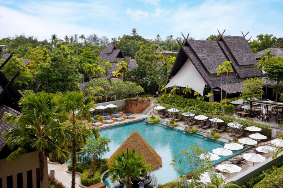 A serene resort featuring traditional-style buildings with steeply sloped roofs surrounded by lush greenery. In the foreground, there is a large, inviting swimming pool bordered by lounge chairs under umbrellas. Tropical trees and plants enhance the tranquil atmosphere of this gem among Phuket attractions.