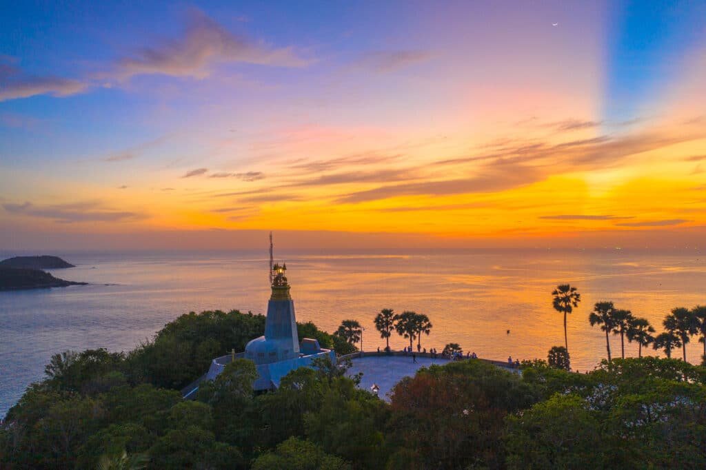 A scenic coastal view at sunset from Laem Phromthep, with a tall, blue structure topped with a golden spire in the foreground. The sky is painted in hues of orange, yellow, and blue with a crescent moon visible. The structure is surrounded by lush greenery and palm trees, with calm ocean waters extending to the horizon.