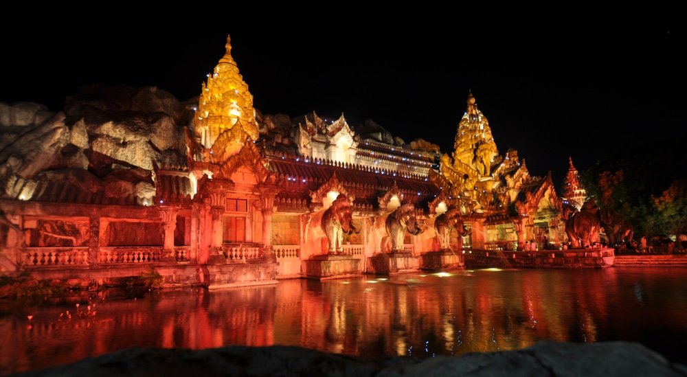 A majestic temple, one of the premier Phuket attractions, is illuminated at night with vibrant yellow and red lights reflecting on a calm body of water in front. The ornate structure features elaborate carvings and statues, including several elephant figures. The dark sky provides a stark contrast, highlighting the temple's intricate details.