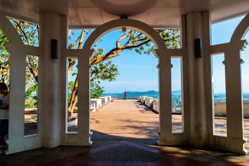 View from inside a structure with white arches, looking out towards a person standing on a scenic overlook. The overlook has railings and benches, offering a panoramic view of the city below, surrounded by greenery and distant mountains under a clear blue sky—truly one of the must-see Phuket attractions.