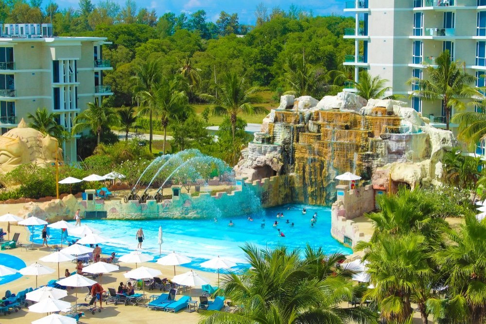 A vibrant resort features a large swimming pool with cascading waterfalls and water jets, reminiscent of top Phuket attractions. Guests swim and relax under sun umbrellas. Surrounding the pool are tall, modern buildings, palm trees, and lush greenery in the background under a clear, blue sky.