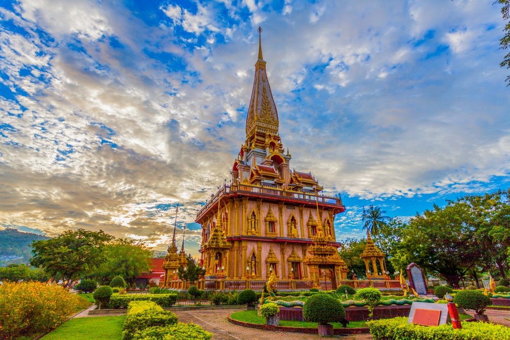 A majestic golden temple with intricate details and a tall spire stands under a vibrant sky with scattered clouds. Lush green shrubs and plants surround the temple, enhancing its grandeur. The setting sun bathes the scene in a warm, radiant light, creating a serene and awe-inspiring atmosphere.