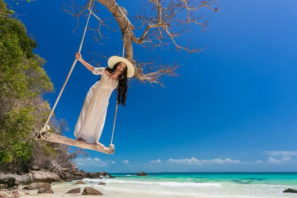 A woman in a long, flowing white dress and a wide-brimmed hat stands on a wooden swing hanging from a bare tree branch by Patong Beach. The sea is calm with turquoise water and clear skies above. The coastline, known for its Patong Beach attractions, is dotted with rocks and trees visible in the background.