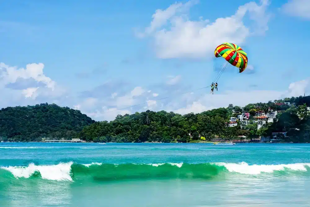 A vibrant beach scene with azure waves crashing on the shore. A parasailor, using a colorful yellow, red, and green parachute, is gliding high above the turquoise water. In the background, a lush green hill dotted with houses hints at Patong Beach attractions. The sky is a bright blue with scattered clouds.