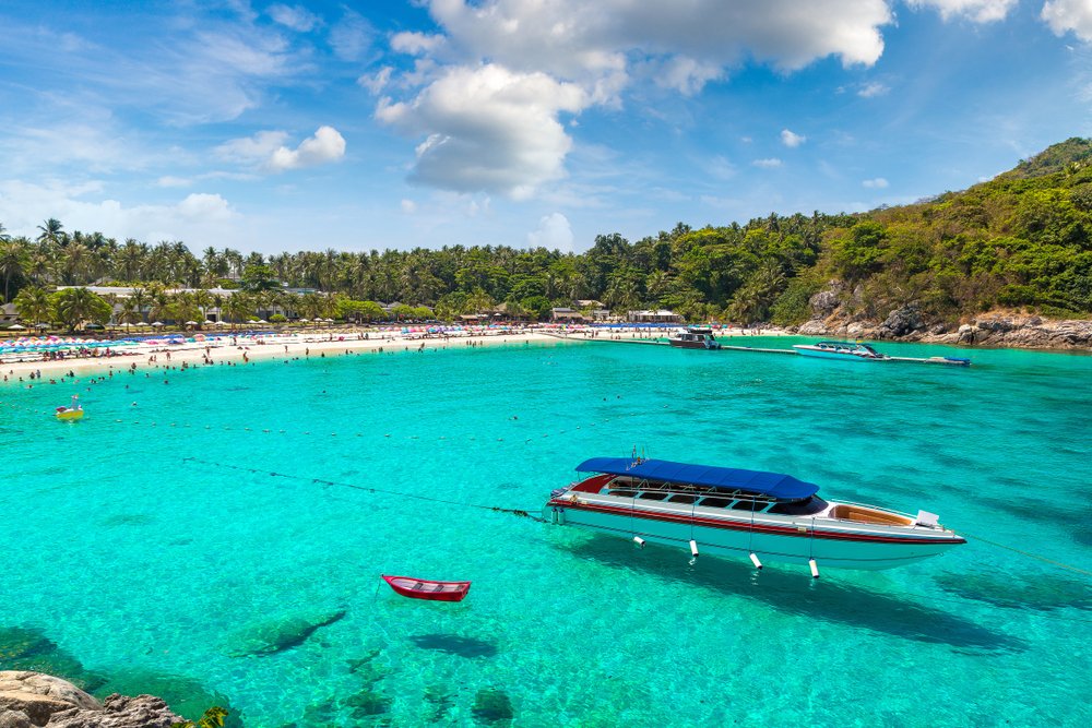 A longtail boat and a red rowboat float on clear turquoise waters near a tropical island. White sandy beaches are lined with people, palm trees, and beach umbrellas. The sky is partly cloudy, and lush green vegetation covers the hills in the background, showcasing highlights from the Phuket travel guide.