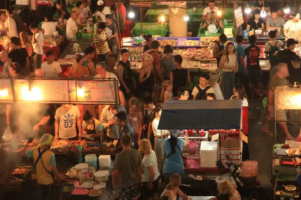 A bustling night market scene with many people shopping and dining at various stalls, exemplifying Patong Beach attractions. The stalls display colorful arrays of goods and food, warmly lit with hanging lights. The lively atmosphere showcases numerous people interacting and moving around the crowded market.