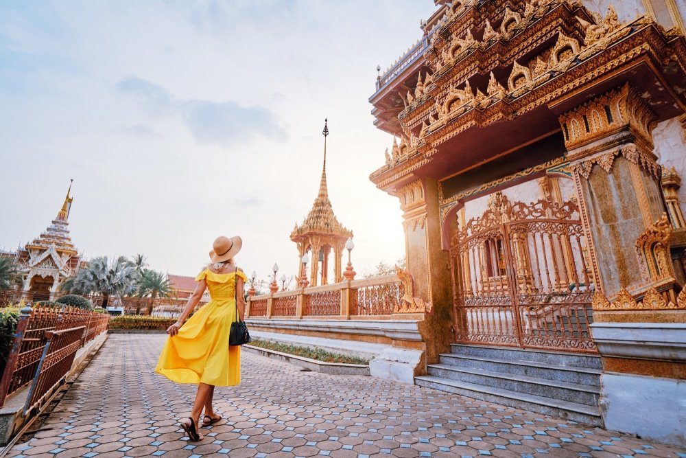 A woman in a flowing yellow dress and sun hat stands on a paved walkway, gazing at a grand, ornate temple adorned with gold decorations. The temple features intricate architecture with spires and carvings. The sky is partly cloudy, and another smaller temple is visible in the background.