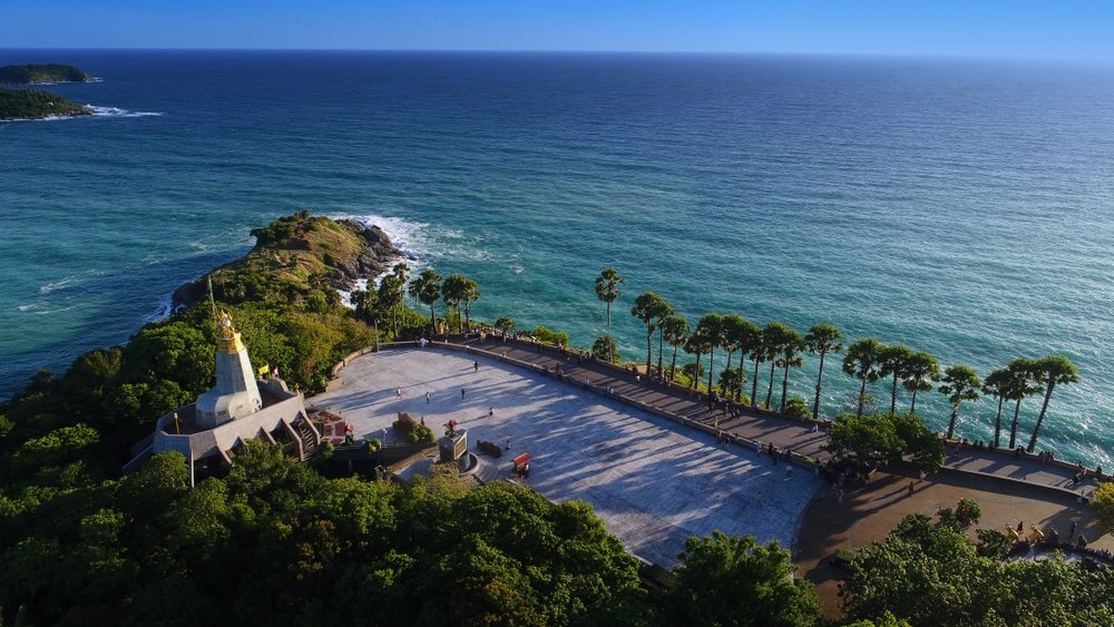 Aerial view of Promthep Cape in Phuket, Thailand. A white lighthouse with a golden dome is on a small hill at the tip of the cape, surrounded by lush greenery. A paved viewing area with people and several palm trees overlooks the vast blue ocean with the horizon in the distance—a must-see in any Phuket travel guide.