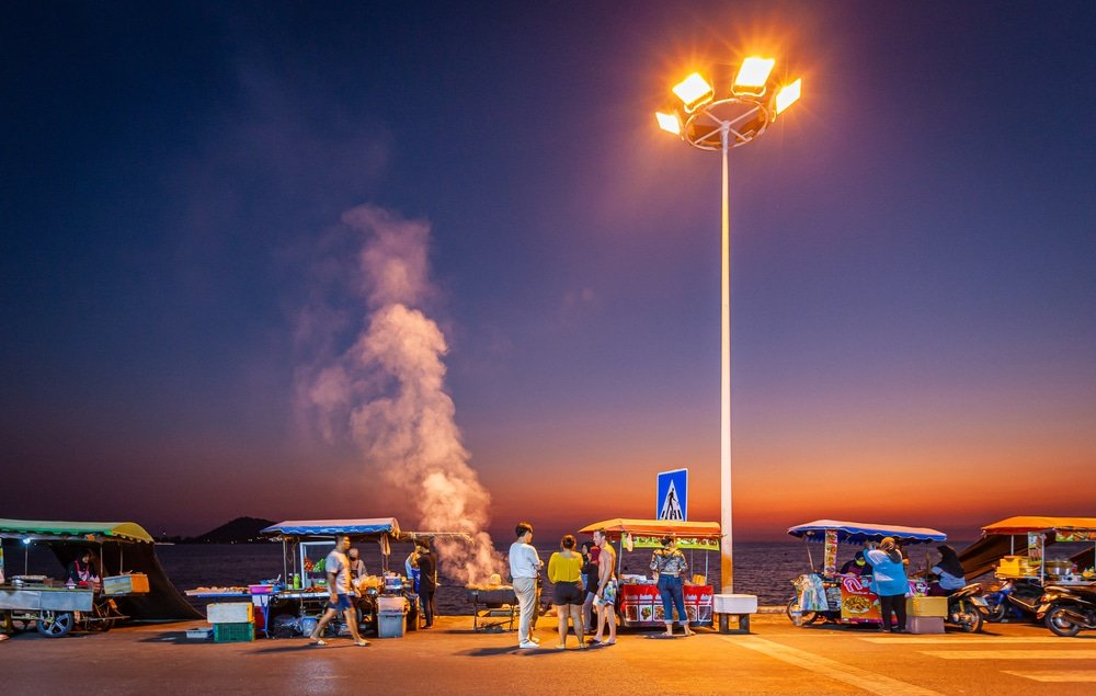 A sunset scene by the coastline of Patong Beach features colorful street food stalls with people gathered around, enjoying one of the popular Patong Beach attractions. A street lamp illuminates the area, and white smoke rises from one of the stalls. The ocean and mountains are visible in the background under a deep orange and purple sky.