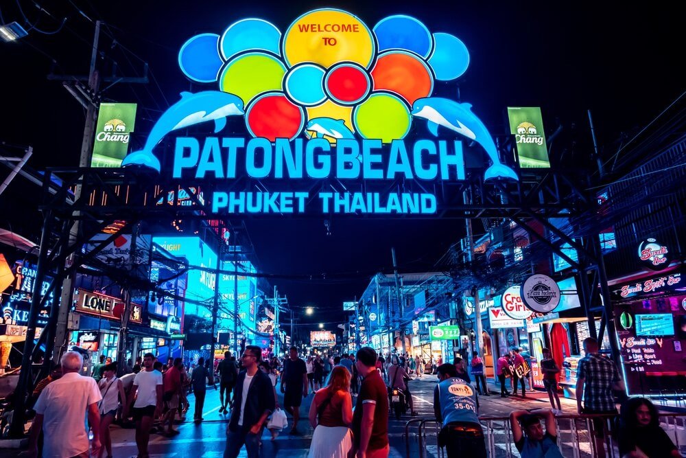 A vibrant, neon-lit street in Patong Beach, Phuket, Thailand, bustling with people at night. A large, brightly lit sign overhead features colorful balloons and two dolphins, welcoming visitors. Numerous bars, restaurants, and shops line the street with radiant neon lights—essential stops in any Phuket travel guide.