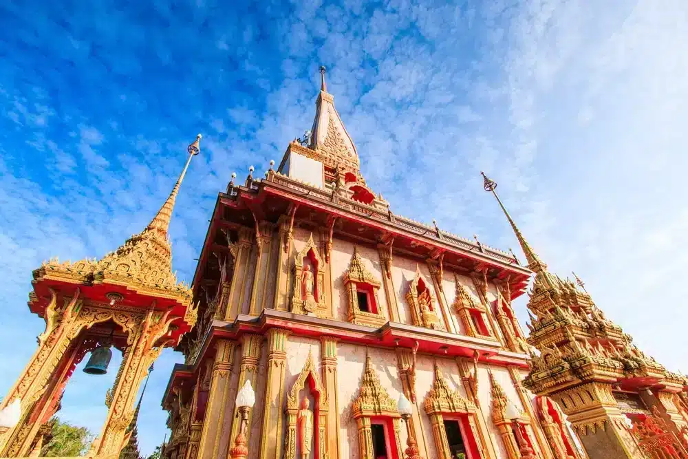 The image shows Wat Chalong Phuket, a Buddhist temple in Thailand. The temple has intricate golden decorations, red accents, and several spires. The sky is bright blue with scattered clouds, creating a vibrant backdrop for the ornate architecture of Chalong Temple.