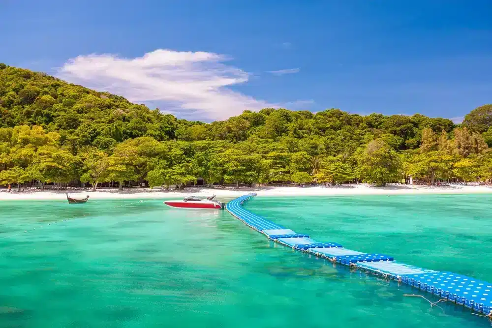 A scenic beach scene with vibrant turquoise water and a floating blue walkway leading to the white sandy shore lined with lush green trees. A motorboat and a traditional boat are anchored near the shore against a backdrop of clear blue sky with a few scattered clouds, making it one of the best places to visit in Patong.