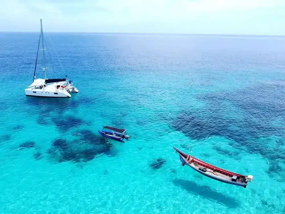 Aerial view of a clear, turquoise sea with three boats floating off Patong Beach. On the left is a white catamaran with people on board, in the middle is a small blue motorboat, and on the right is a long, narrow traditional wooden boat. The water beneath shows dark patches of underwater reefs.