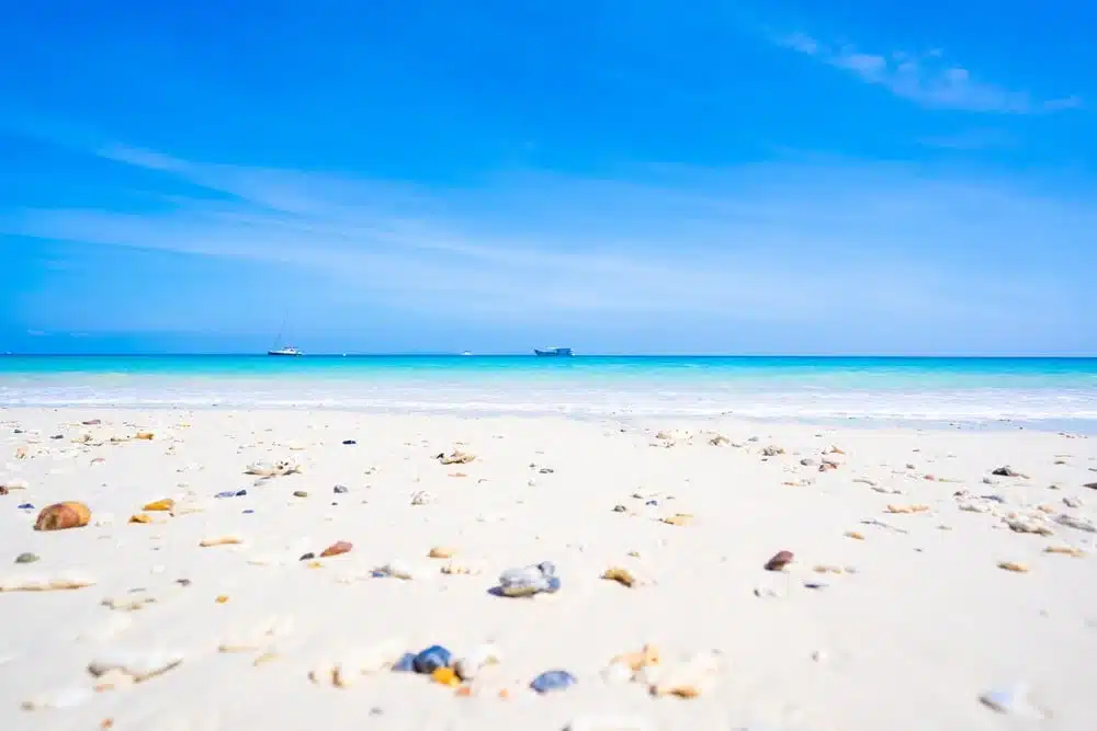 A serene Patong Beach scene with a clear blue sky and turquoise ocean waters. Smooth white sandy shore in the foreground with scattered pebbles and shells. Two boats are visible in the distance on the horizon, making it one of the best places to visit in Patong for a sense of calm and tranquility.