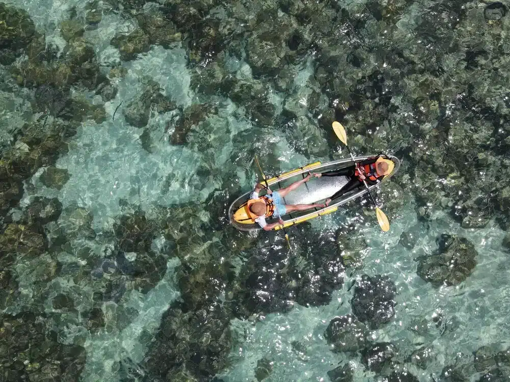 Aerial view of a person in a dark-colored kayak, paddling through clear turquoise waters with rocky formations visible underneath near Patong Beach attractions. The person wears a hat and is seated in the kayak, which also carries a large fish on board. The sun casts light reflections on the water’s surface.