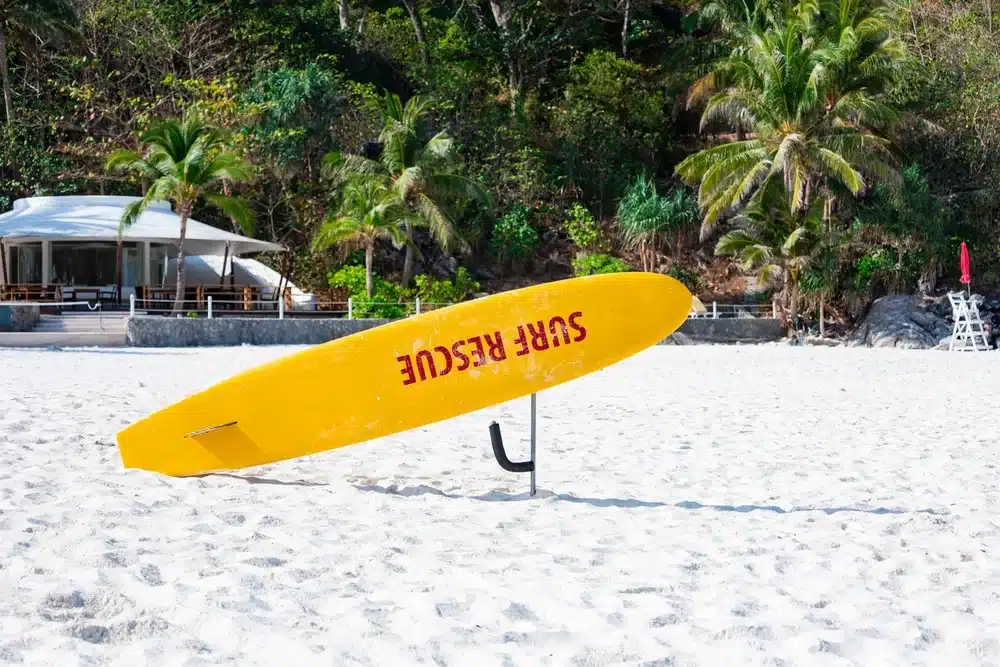 A yellow surfboard with the text "SURF RESCUE" in red stands upright on the white sandy beach, framed by lush green and palm trees. In the distance, a shelter with a roof and wooden deck, along with a white lifeguard chair, hints at why this spot is among the best places to visit in Patong.