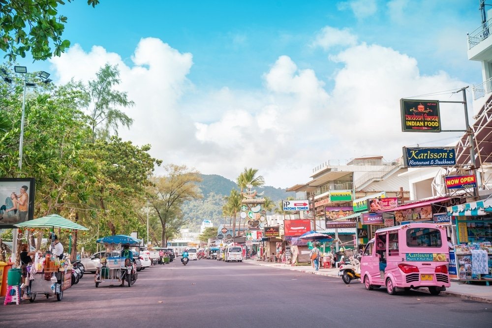 A vibrant street scene in Patong with various shops and restaurants on both sides. Signs for businesses, including an Indian food restaurant and a visible pharmacy, line the street. Several vehicles, including a pink tuk-tuk and motorcycles, are on the road. With mountains and a partly cloudy sky in the background, it’s one of the best places to visit in Patong.