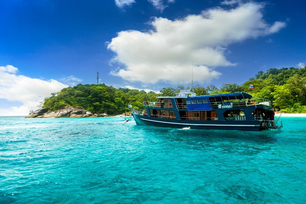 A large blue boat floats on clear turquoise water near a lush, green island under a bright blue sky with scattered clouds. The island features dense vegetation and rocky shores. The boat is equipped with various supplies and gear, suggesting potential recreational or fishing activities—perfect for Phuket travel guide recommendations.