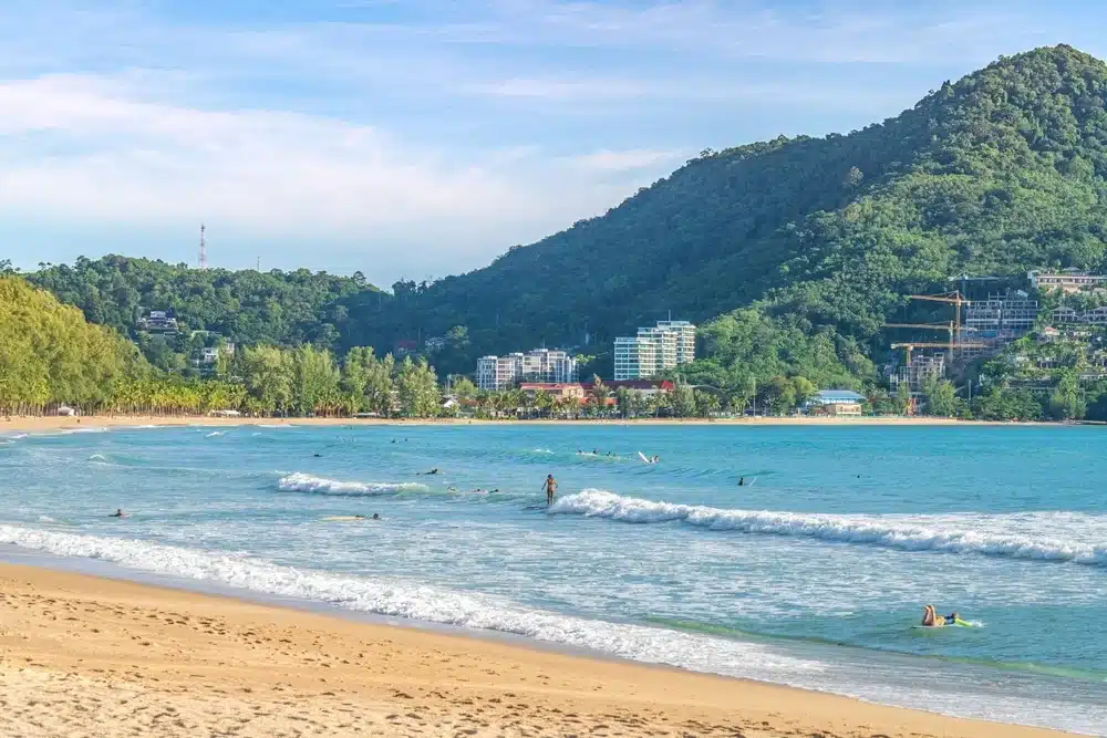 A tranquil beach scene features golden sand and gentle blue waves under a clear sky. The shoreline curves gracefully, leading to a lush, tree-covered hill with a few buildings in the distance. People are enjoying water activities, creating a relaxed and inviting atmosphere—one of the best places to visit in Patong.