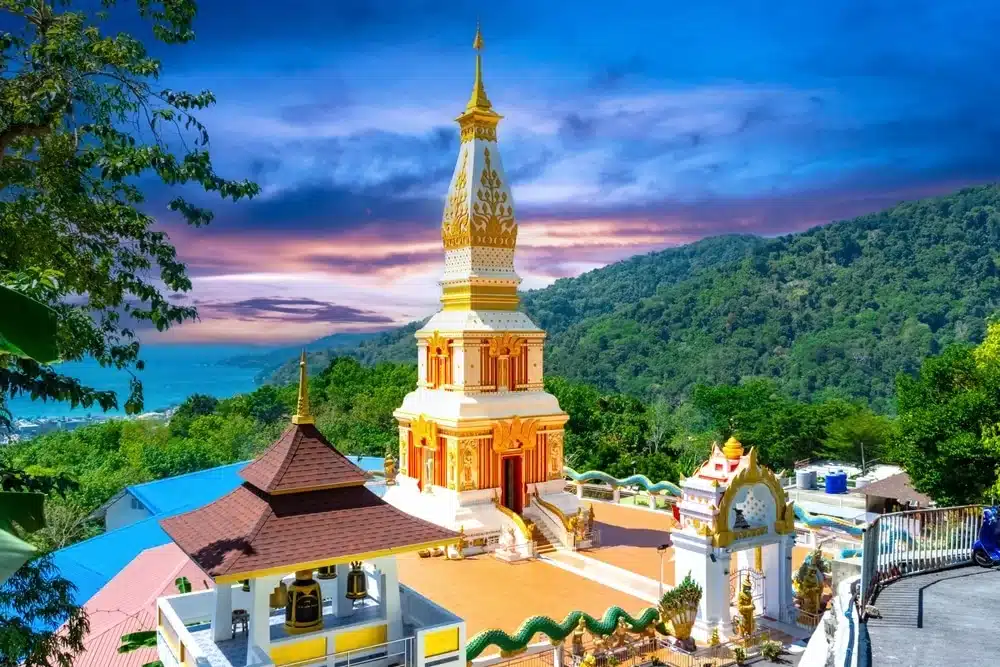 A stunning Buddhist temple with a golden and white stupa stands prominently, surrounded by lush greenery and mountains. A vibrant sunset sky with hues of blue, purple, and orange provides a breathtaking backdrop. Discover one of the best places to visit in Patong as the ornate staircase leads up to the entrance.