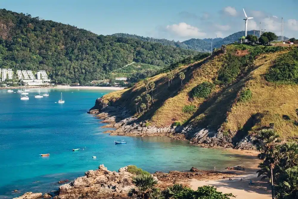 A scenic coastal landscape showcases a lush, green hill with a wind turbine on top, overlooking a clear blue bay dotted with anchored boats. The shoreline features rocky terrain and a small sandy beach. In the background, forested hills and some residential buildings can be seen—a scene reminiscent of Laem Phromthep Sunset Viewpoint.