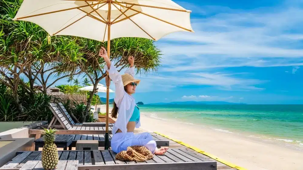 A person in a sun hat and sunglasses lounges on a wooden deck chair under a large white umbrella, enjoying one of the best places to visit in Patong. The person is stretching their arms upwards. The sandy beach, turquoise water, and clear blue sky create a serene, tropical backdrop. There is a pineapple and straw bag nearby.