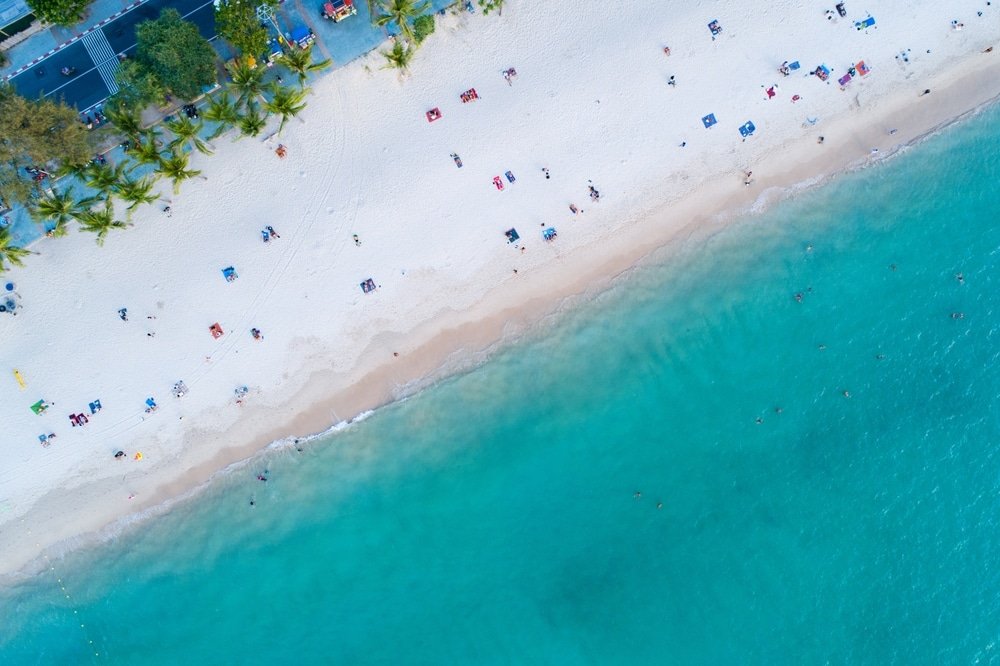 Aerial view of a sandy beach with turquoise waters, one of the top Phuket attractions. The beach is lined with palm trees and has scattered sunbathers, umbrellas, and towels. The ocean transitions from light to deep blue. People are swimming in the water while others relax on the sand or under shaded areas near the trees.