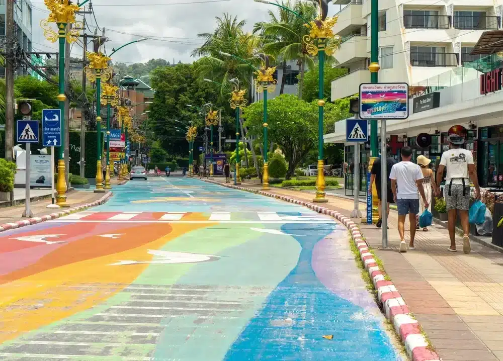 A vibrant street scene features a wide road painted with colorful patterns, including a white zebra crossing. Golden decorative streetlights line the street. On the right, two people walk near modern buildings and green trees, reminiscent of Patong Beach attractions. Various signs and shops are visible in the background.