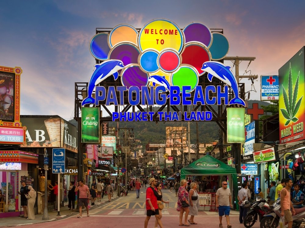 A bustling street at Patong Beach, Phuket, Thailand, lit with vibrant neon signs and a prominent "Welcome to Patong Beach" sign adorned with dolphins. Tourists and locals meander among shops, bars, and cafes. The background features hills under a twilight sky—one of the best places to visit in Patong.