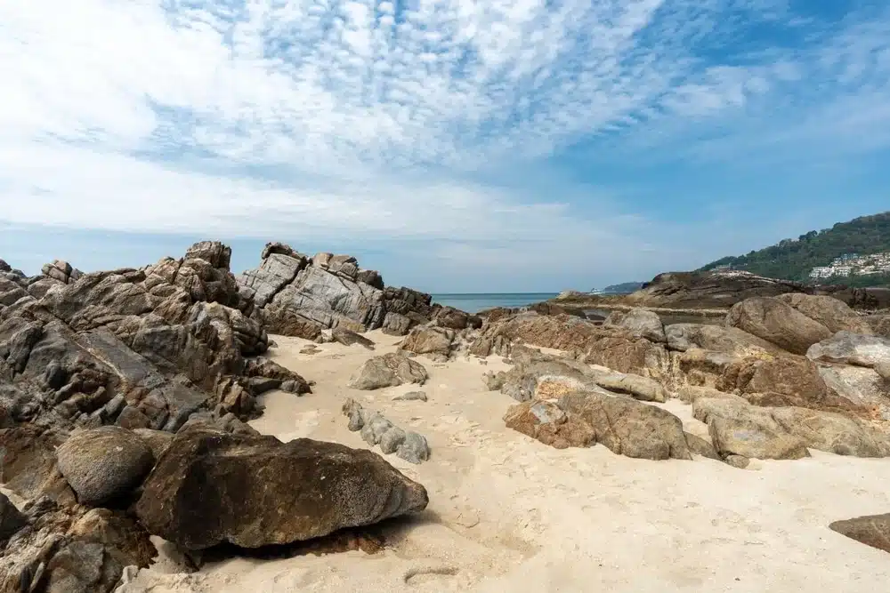 A coastal scene featuring a sandy beach interspersed with large rocky formations, reminiscent of Patong Beach attractions. The sky is partly cloudy with patches of blue and streaks of white clouds, while distant hills covered in greenery extend towards the sea, creating a serene, natural landscape.