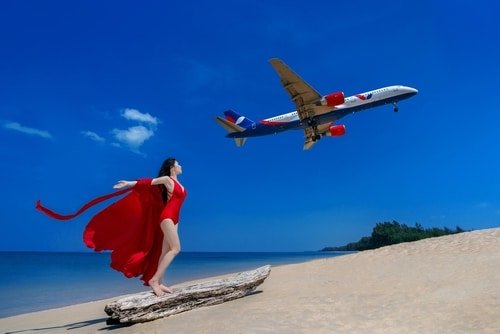 A woman in a red dress with a long flowing cape stands on a piece of driftwood at the beach, embodying the allure of Phuket attractions. She poses with arms outstretched, gazing at an airplane flying overhead against a clear blue sky. The ocean is calm, and the sandy beach extends into the distance with some greenery visible.