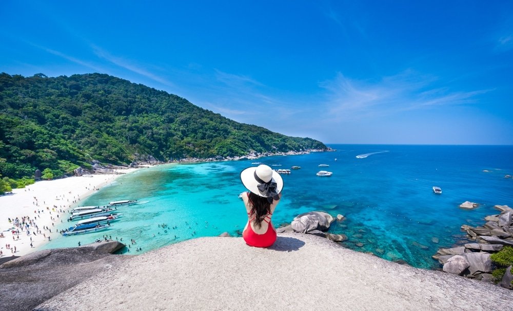 A woman in a red swimsuit and wide-brimmed hat sits on a large rock, overlooking a picturesque beach with clear turquoise water. Several boats are anchored near the shore, while sunbathers populate the white sandy beach. A lush, green, hilly landscape forms the backdrop under Phuket's clear blue sky.