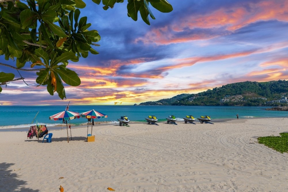 A tropical beach at sunset features a vibrant sky with hues of pink, purple, and orange. The sandy shore is dotted with colorful umbrellas and lounge chairs on the left. A row of jet skis is lined up near the water. Lush green hills are visible in the background, with calm blue waters in the foreground—perfect for any Phuket travel guide.