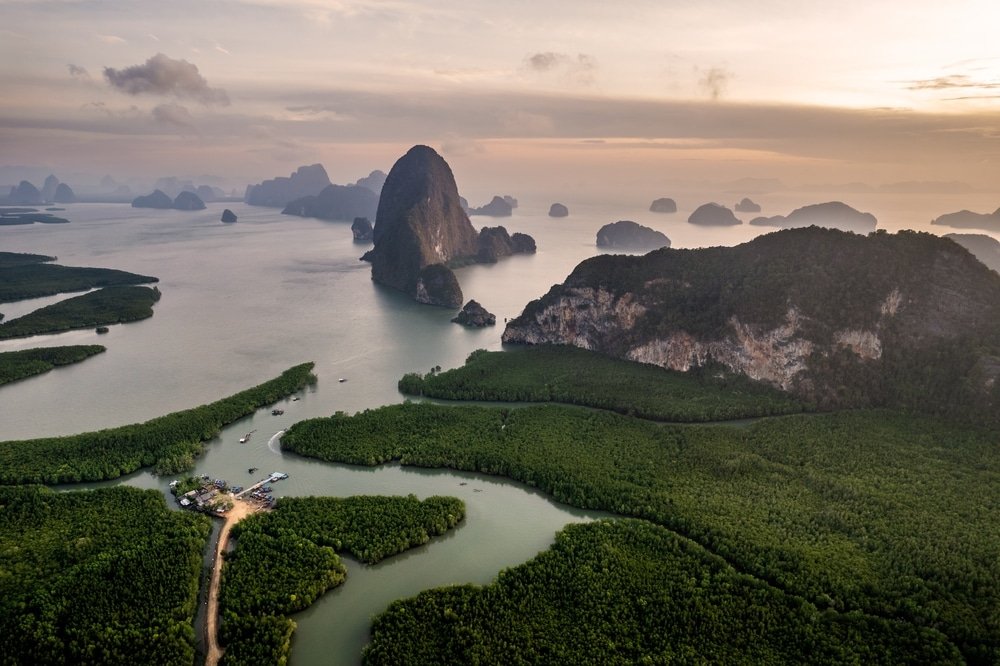 Aerial view of Phang Nga Bay in Thailand at sunset. The bay, a highlight in many Phuket travel guides, features scattered limestone karsts and islets rising from calm, reflective waters. The coastline is lined with dense mangroves, and a small settlement with boats is visible near the shore. Soft, pink-tinted clouds fill the sky.