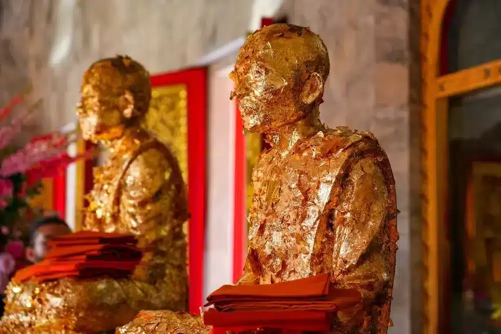 Two intricately gilded Buddha statues sit in a serene temple setting, one of the best places to visit in Patong. Each statue is adorned with gold leaf and dressed in traditional monk robes. The background features red and gold accents and an arched doorway, enhancing the tranquil atmosphere. Rolls of orange fabric are placed on the statues' laps.