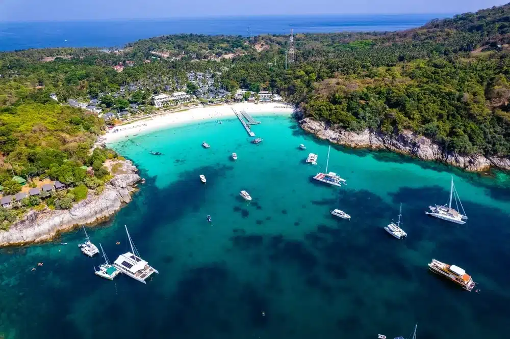 Aerial view of a scenic bay with turquoise waters, surrounded by lush green hills. Several sailboats and yachts are scattered in the water, with a sandy beach and Patong Beach attractions visible in the background. The clear, sunny sky enhances the vibrant colors of the landscape.