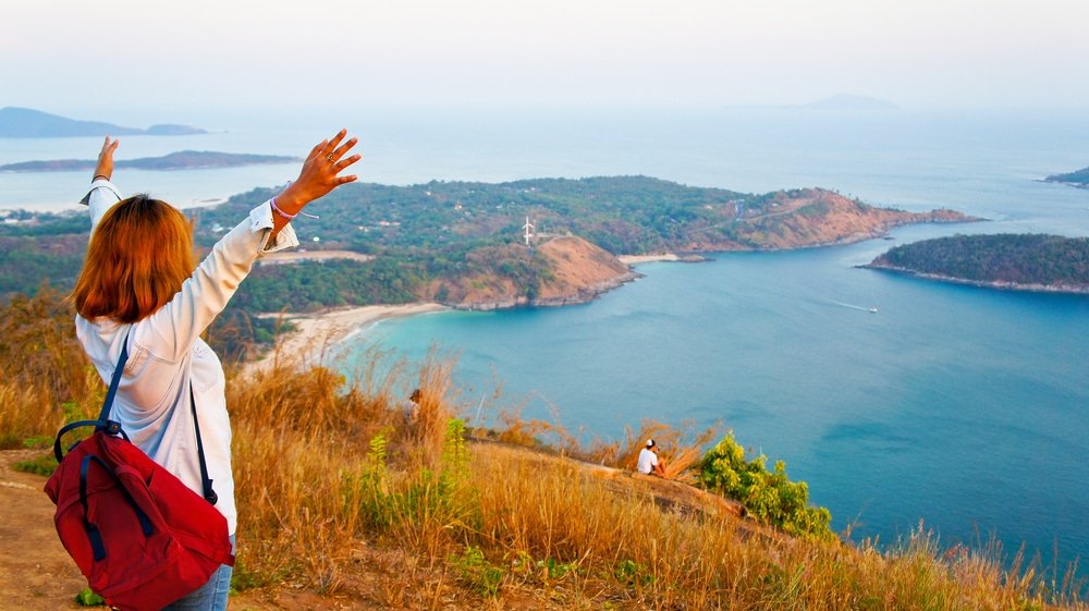 A person with red hair wearing a light-colored shirt and a red backpack raises both arms while standing on a grassy cliff overlooking a vibrant, blue sea with lush greenery and rocky shores—a perfect spot featured in many Phuket travel guides. Another person sits on the edge farther down, taking in the scenic view.