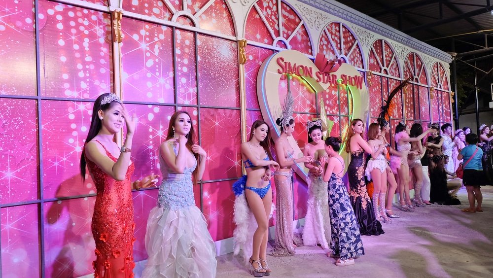 A group of performers stand in a row in front of a pink backdrop with sparkly patterns and the sign "Simon Star Show." They are dressed in elaborate costumes, including gowns, feathered outfits, and bejeweled accessories. This dazzling show is one of the must-see Phuket attractions highlighted in every Phuket travel guide.