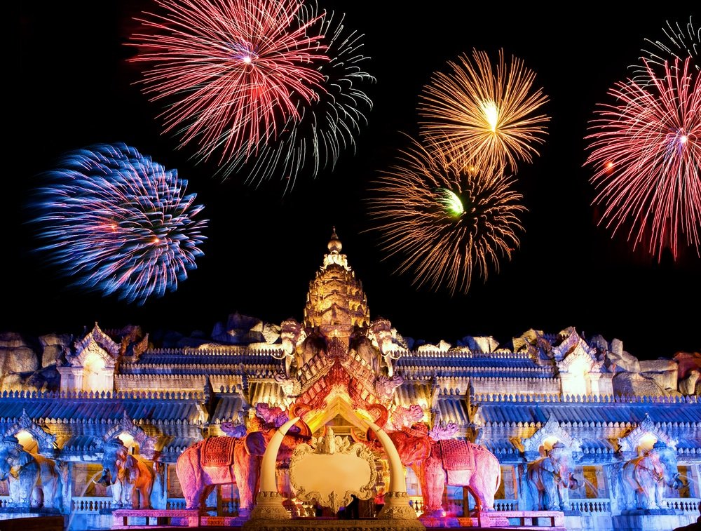 Fireworks light up the night sky above a vibrant, ornately decorated temple, featuring intricate architecture and multiple statues of elephants. The Phuket attractions gem is illuminated in vivid blue, purple, and yellow hues, creating a striking contrast against the dark backdrop.