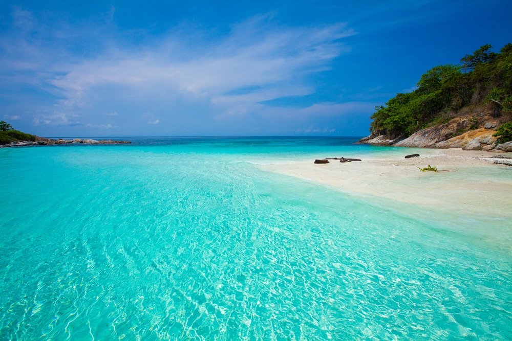 A pristine tropical beach with crystal-clear turquoise waters gently lapping against white sand. The sky is bright blue with scattered clouds. On the right side, there's a small rocky hill covered with lush green vegetation. This serene Phuket travel guide scene showcases the sea extending to the horizon.