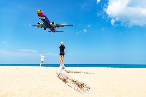 A person stands atop a piece of driftwood on a sandy beach, directly underneath an approaching airplane with purple, orange, and white livery. Another person stands farther back, near the water, shielding their eyes from the sun. This picturesque scene is a highlight in any Phuket travel guide.